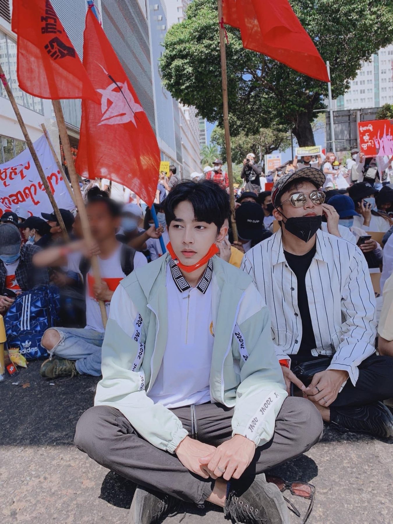 Wai Yan Aung, wearing a red mask under his chin, sits cross-legged on the ground at a crowded rally beneath red flags.