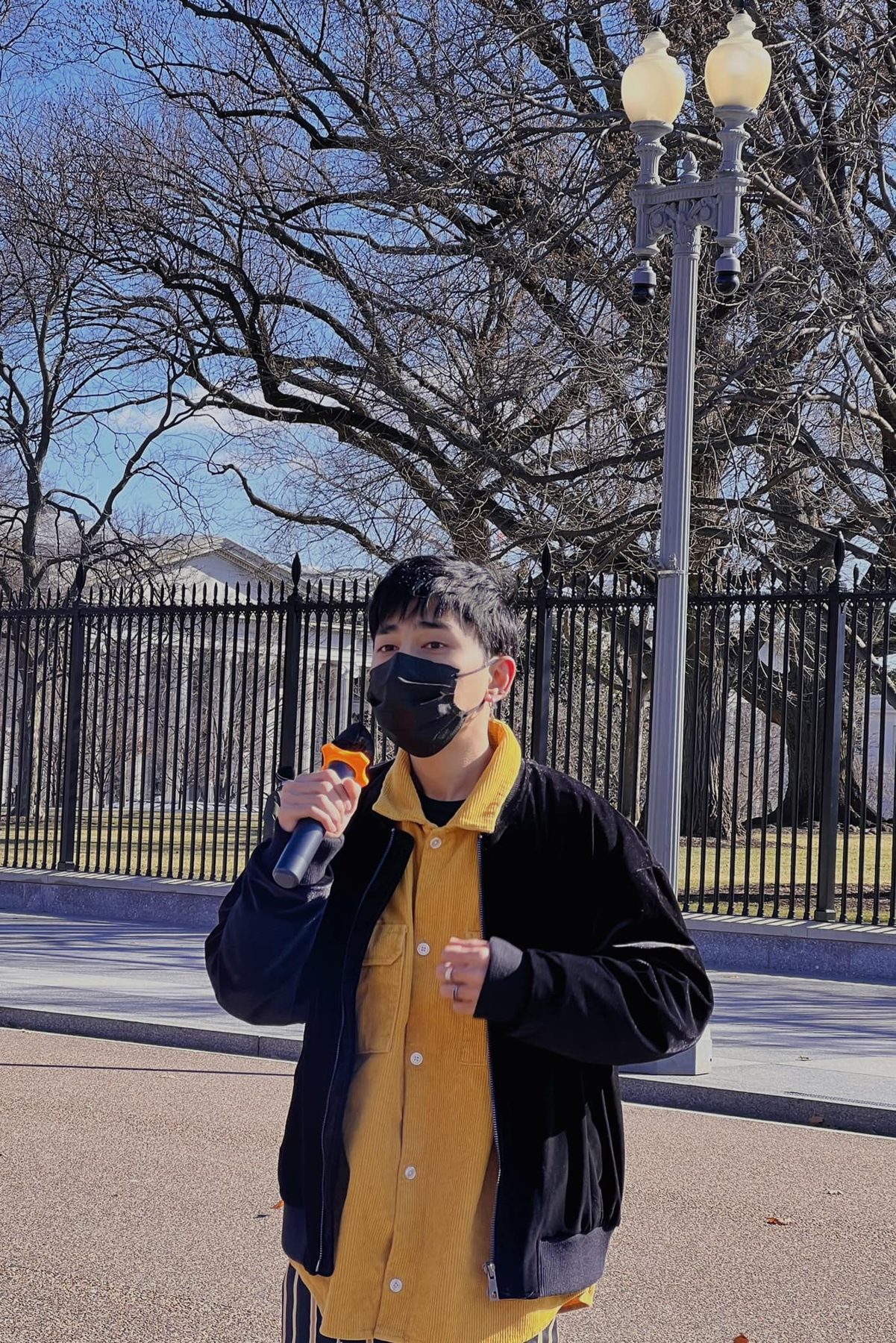Wai Yan Aung, in a black mask and a yellow shirt, speaks into a microphone in front of the White House fence.