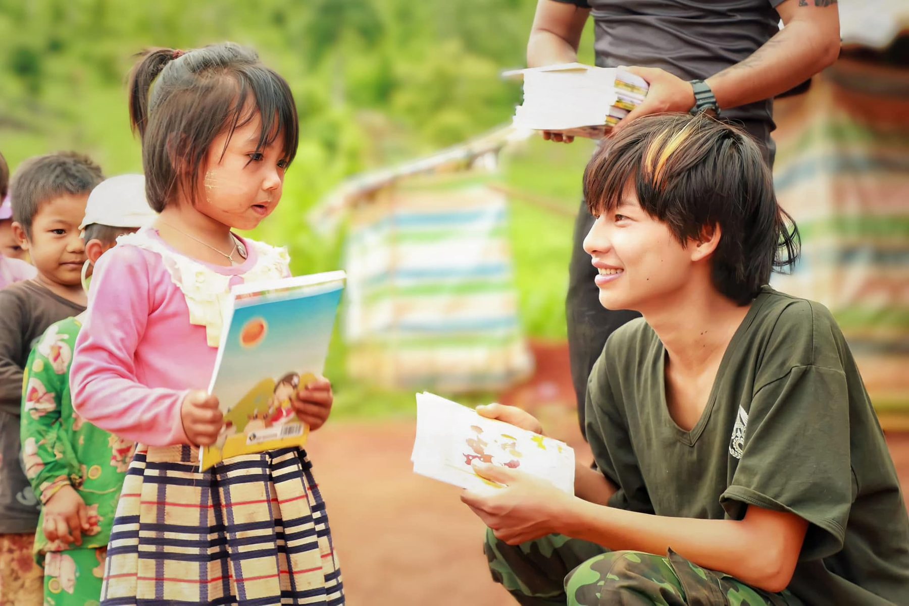 A young girl in a pink jacket holds a new notebook while a smiling volunteer crouches beside her with another notebook, outdoors in a camp.