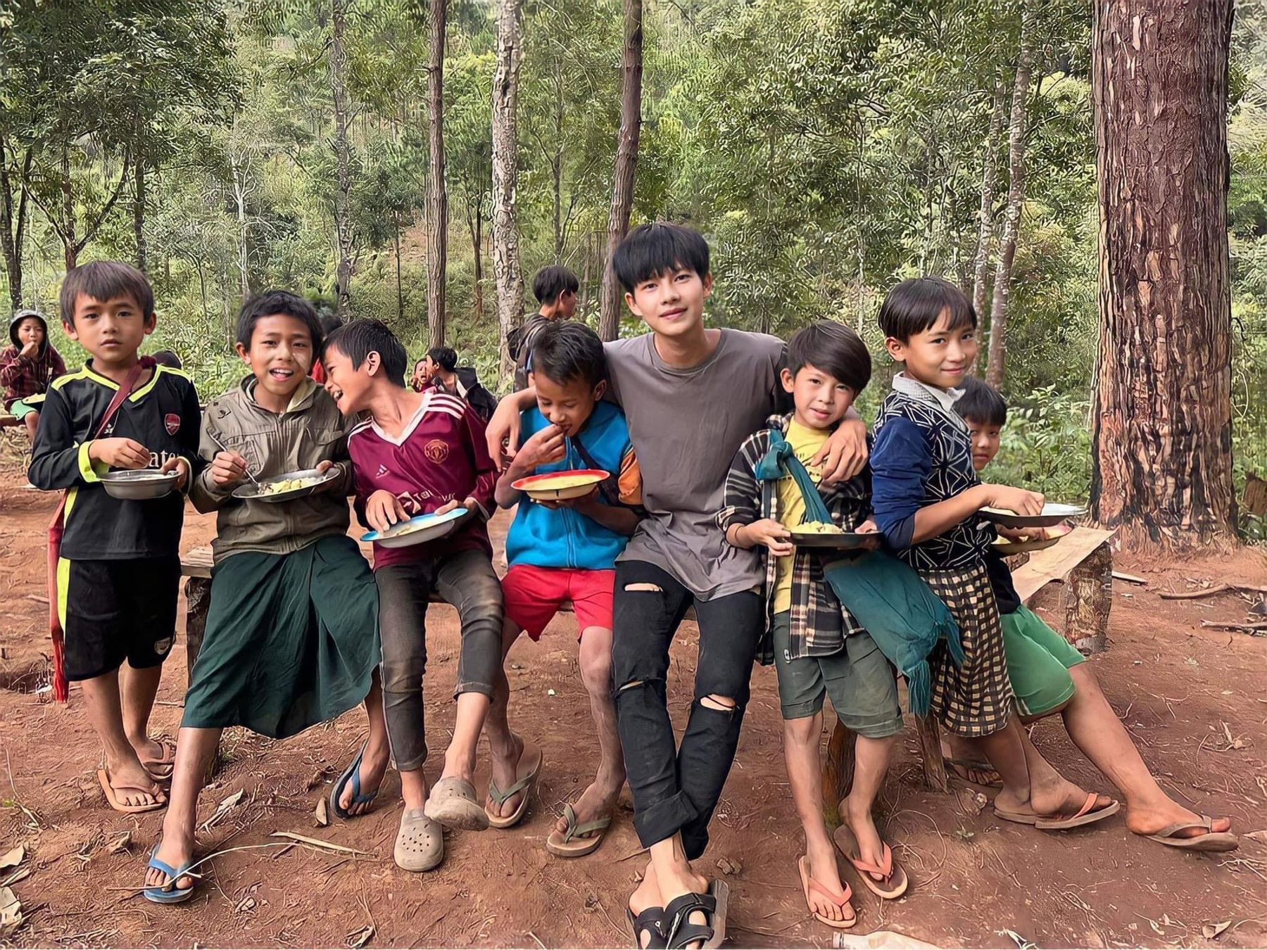 A group of children hold plates of food among tall pine trees.