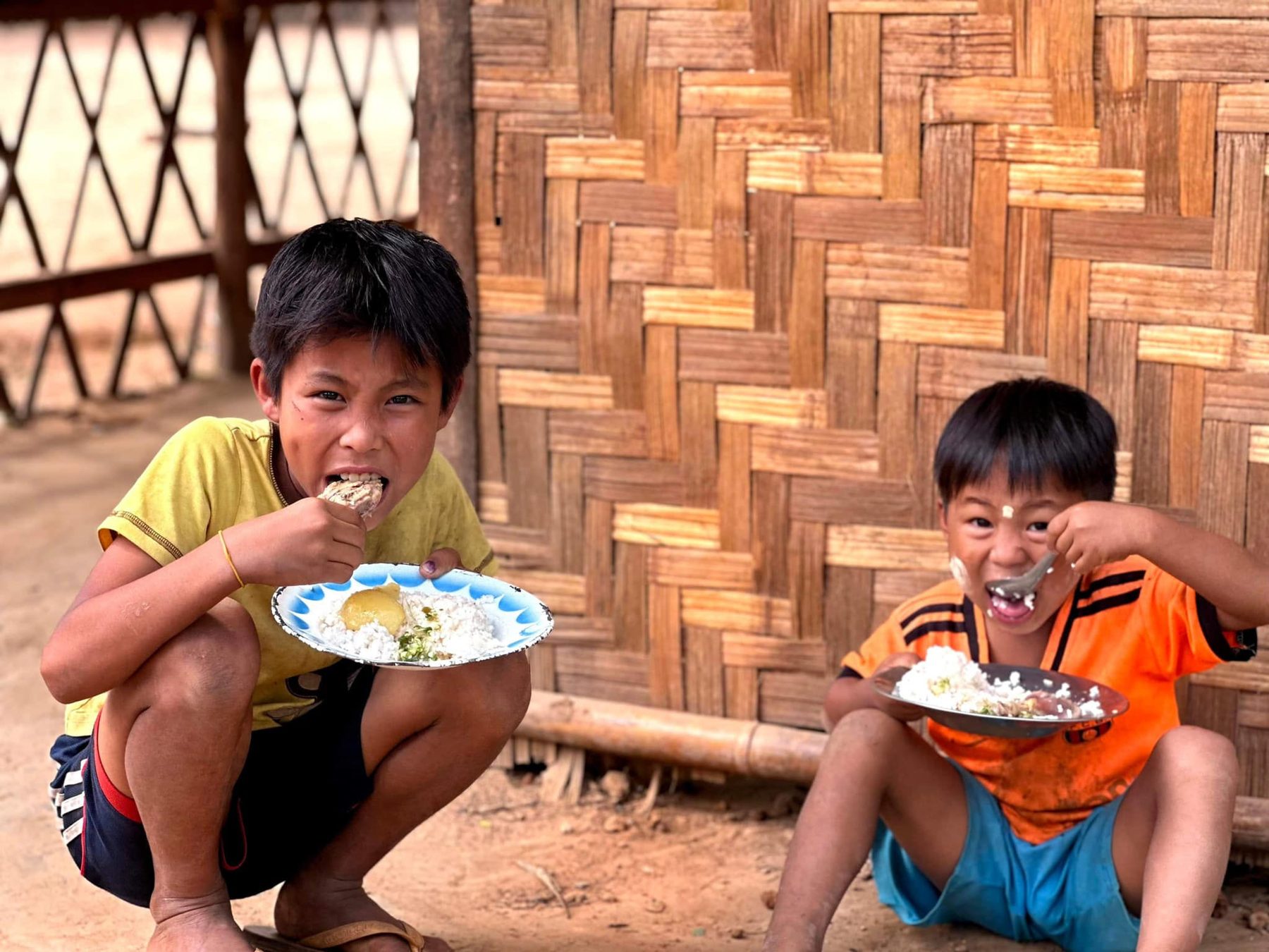 Two boys crouch by a woven bamboo wall, eating rice from plates.