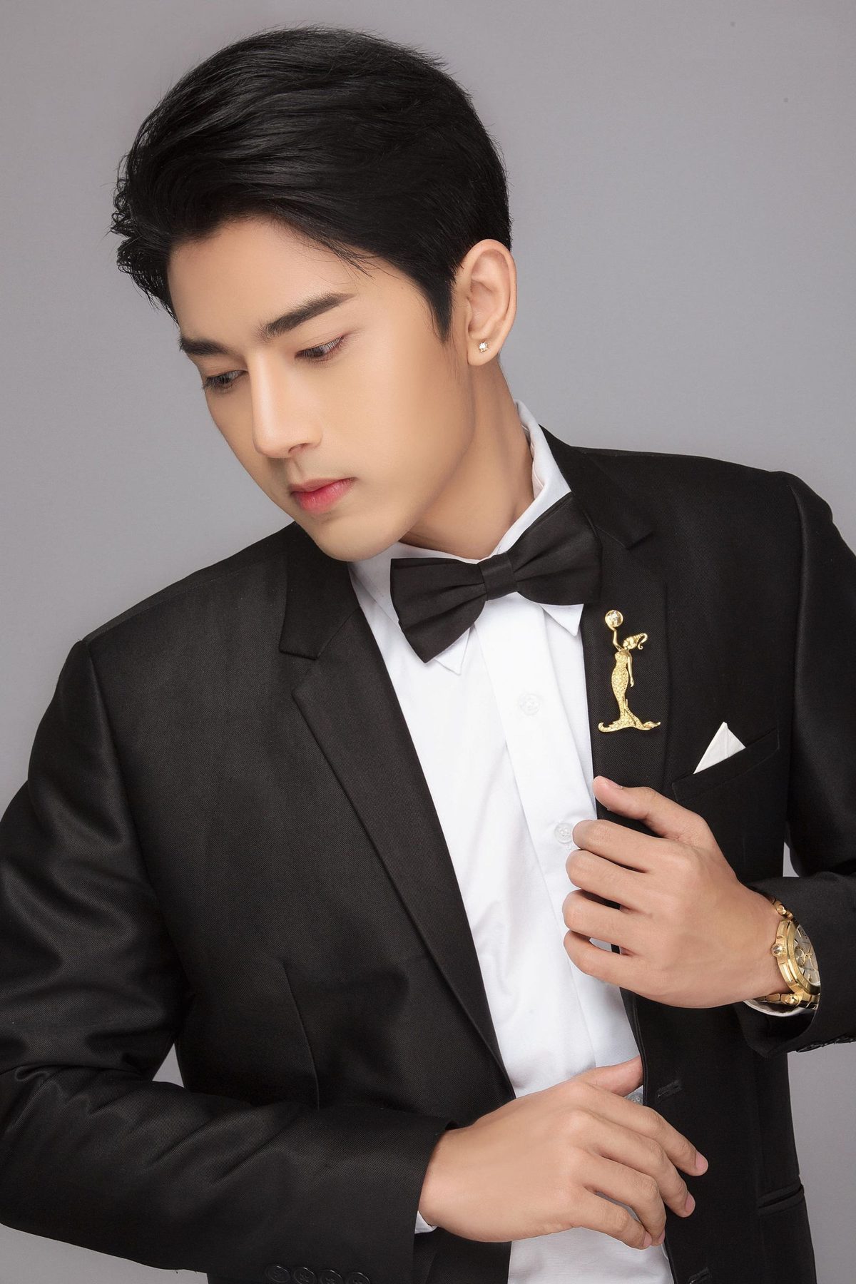 Wai Yan Aung in a black tuxedo with a bow tie and a gold lapel pin, looking down, against a grey background.